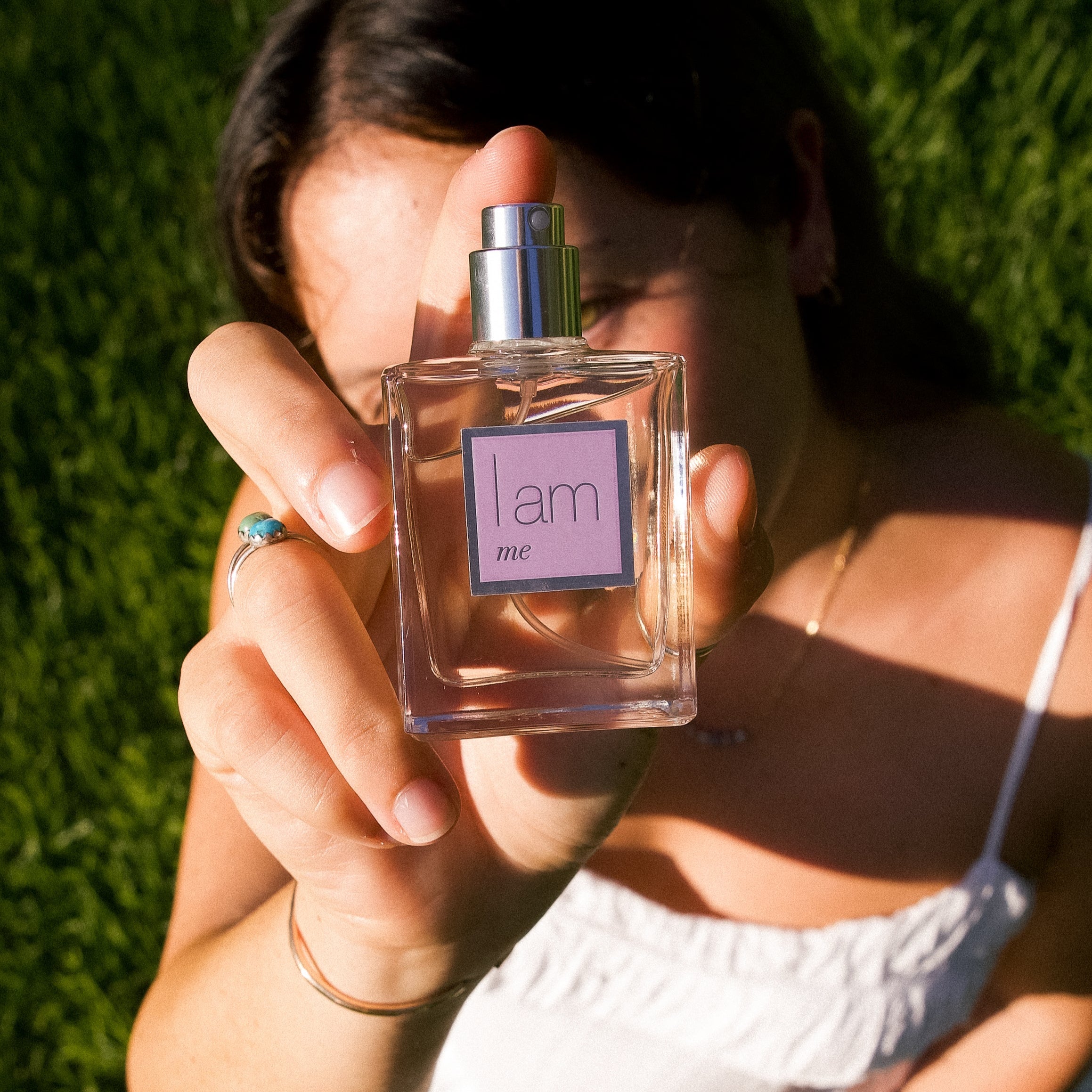 Person holding a perfume bottle labeled 'l am me' against a grassy background