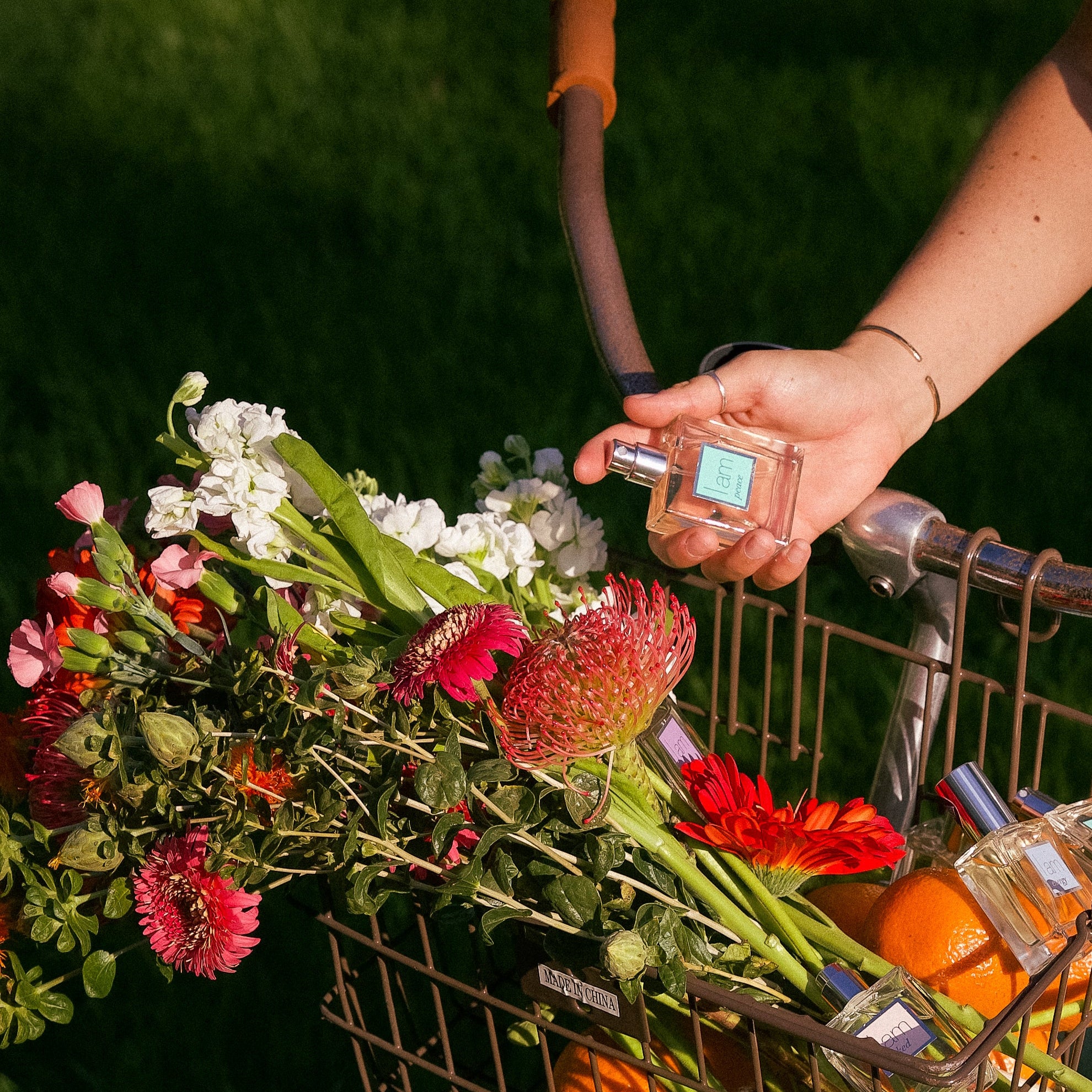 Person holding a bicycle basket filled with flowers and oranges and perfume against a green background
