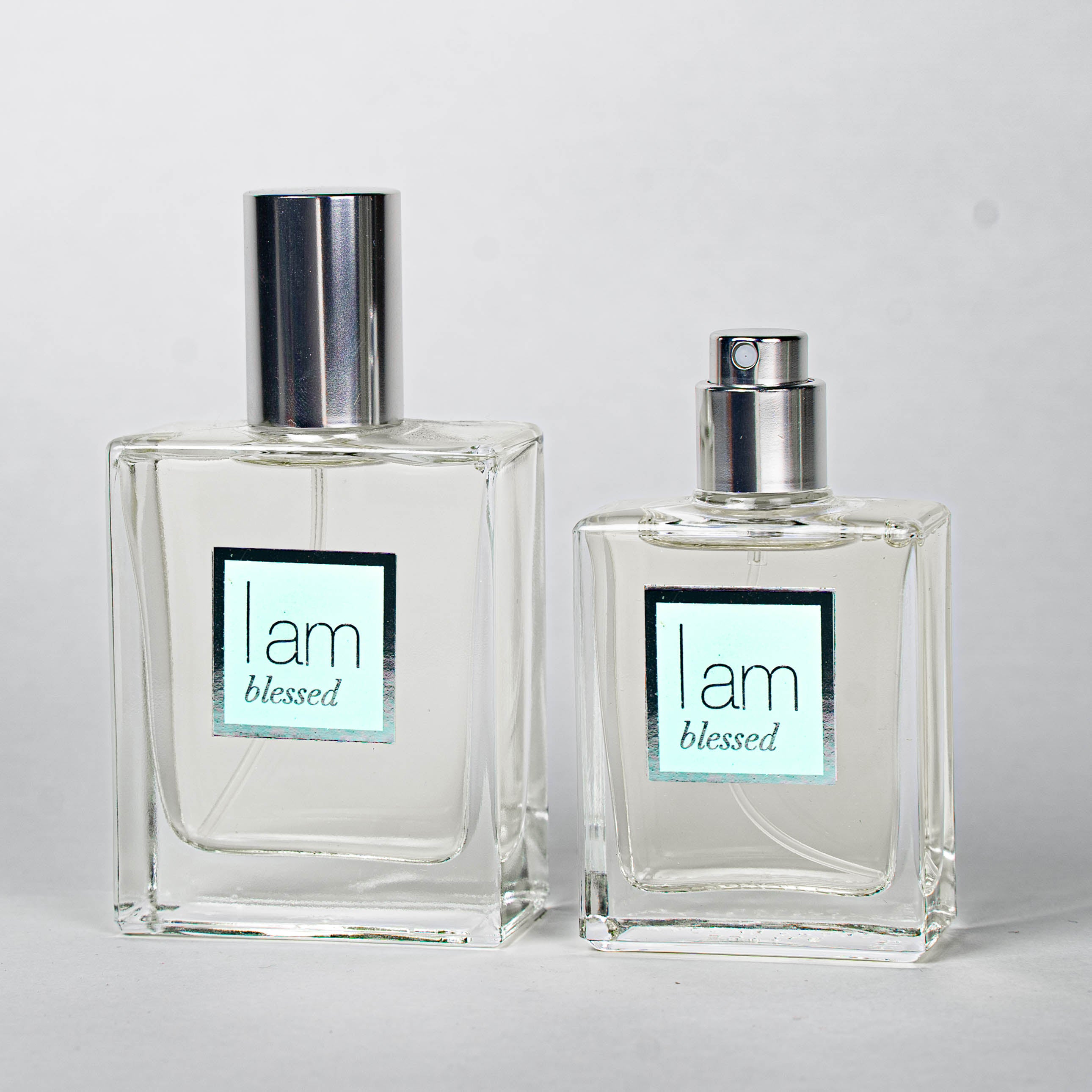 Two clear perfume bottles with silver caps on a light gray background, featuring 'i am blessed' labels.