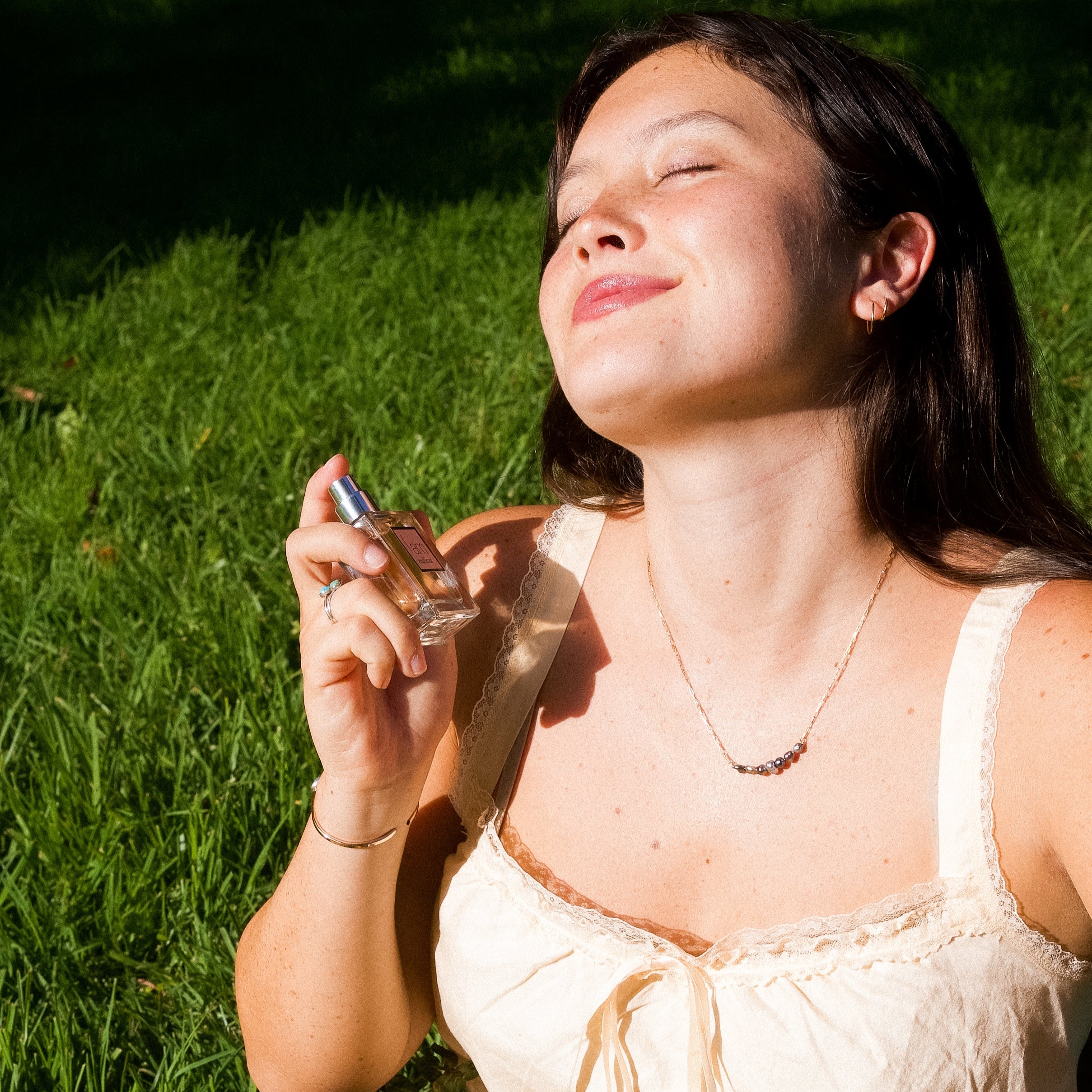 Woman sitting on grass holding flowers and a bottle, with sunlight casting shadows