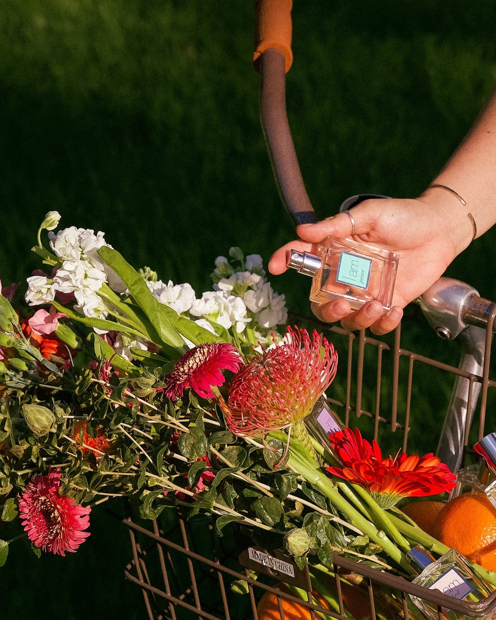 Person holding a bicycle basket filled with flowers and oranges and perfume against a green background